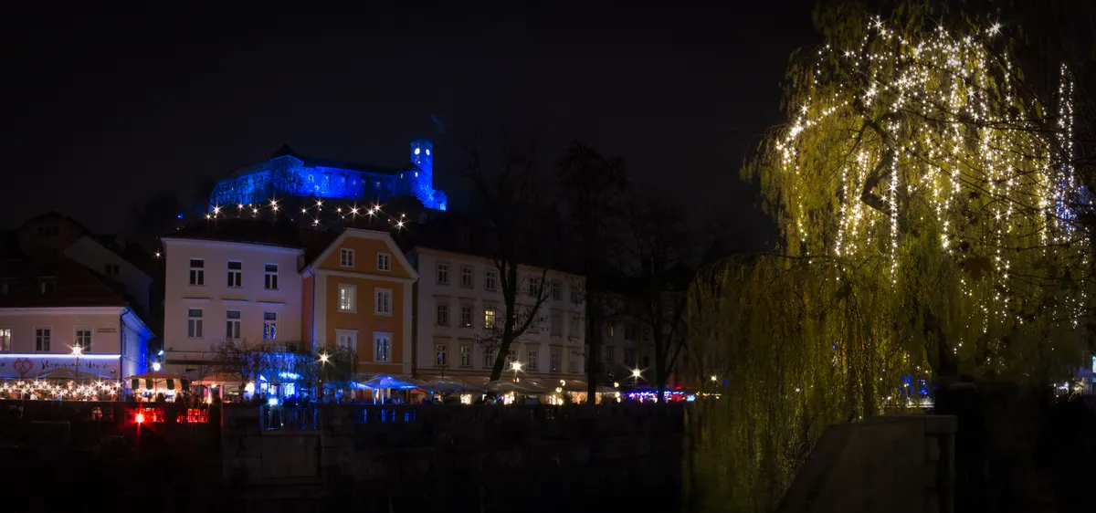 Ljubljanski grad v modri svetlobi, razsvetljena vrba in stojnice ob Ljubljanici v adventu
