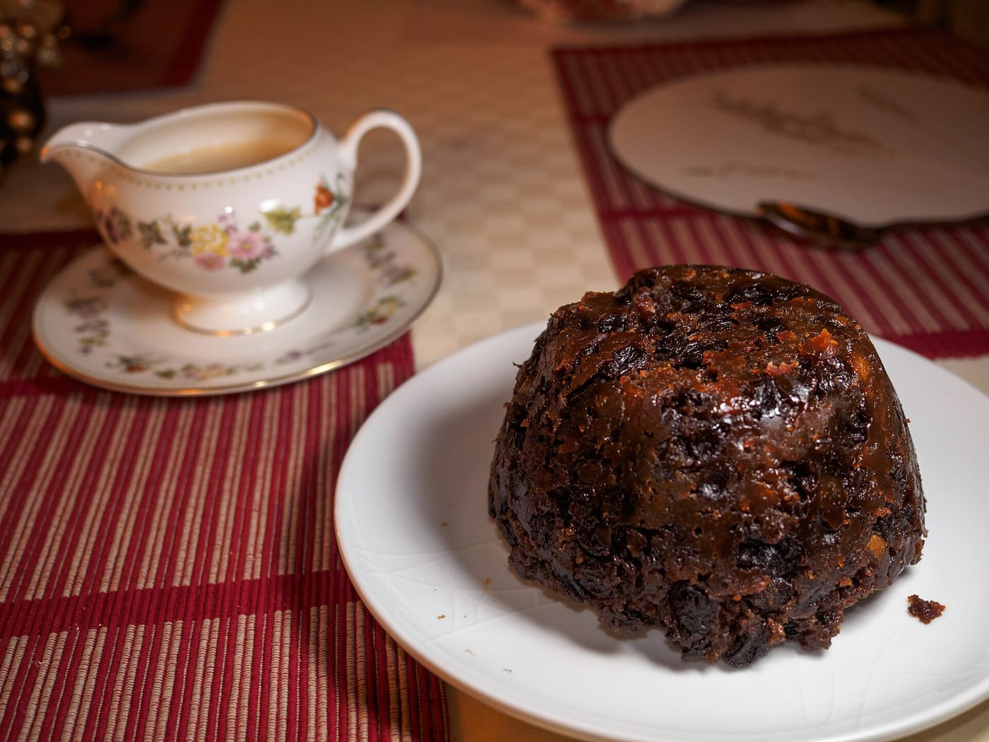 A dark Christmas pudding turned out on a plate, with a jug of custard on a Christmas table
