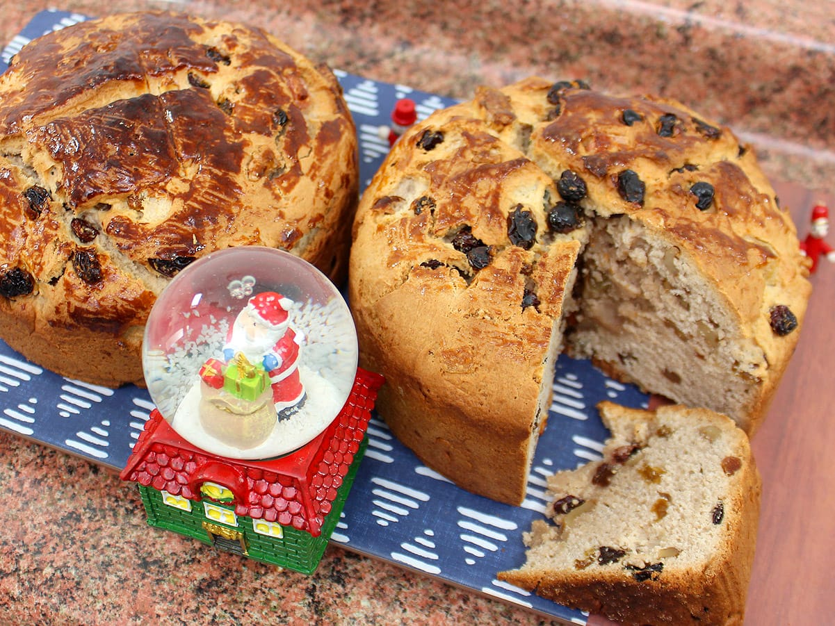 Dos panes de pascua caseros con pasas y frutas, uno cortado, junto a un adorno navideño